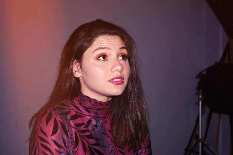 A photo of a non-binary person of color presenting as femme. They have long, straight, dark hair, glitter eye make up, and are wearing a burgundy and black shirt. They are looking to the side and camera equipment can be seen behind them.