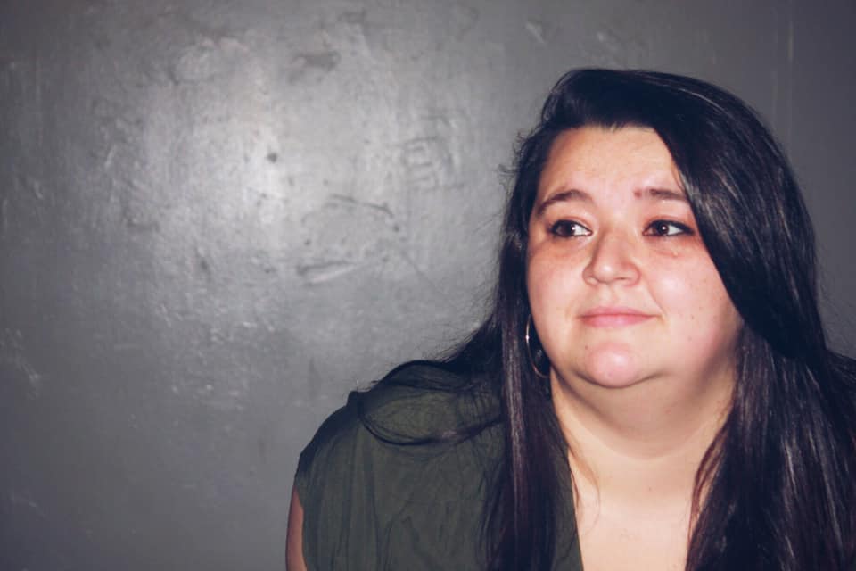 A photo of a white woman with long, dark, straight hair and a green dress. She is positioned on the right side of the photo and is looking to the left.