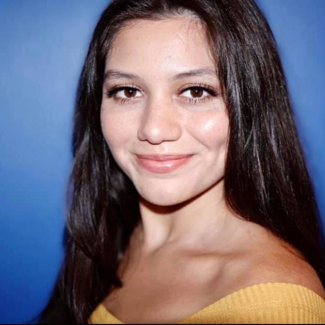 Sam, a femme presenting Latinè person with long brown hair. They are wearing a yellow mustard top and are smiling towards the camera with a blue background.