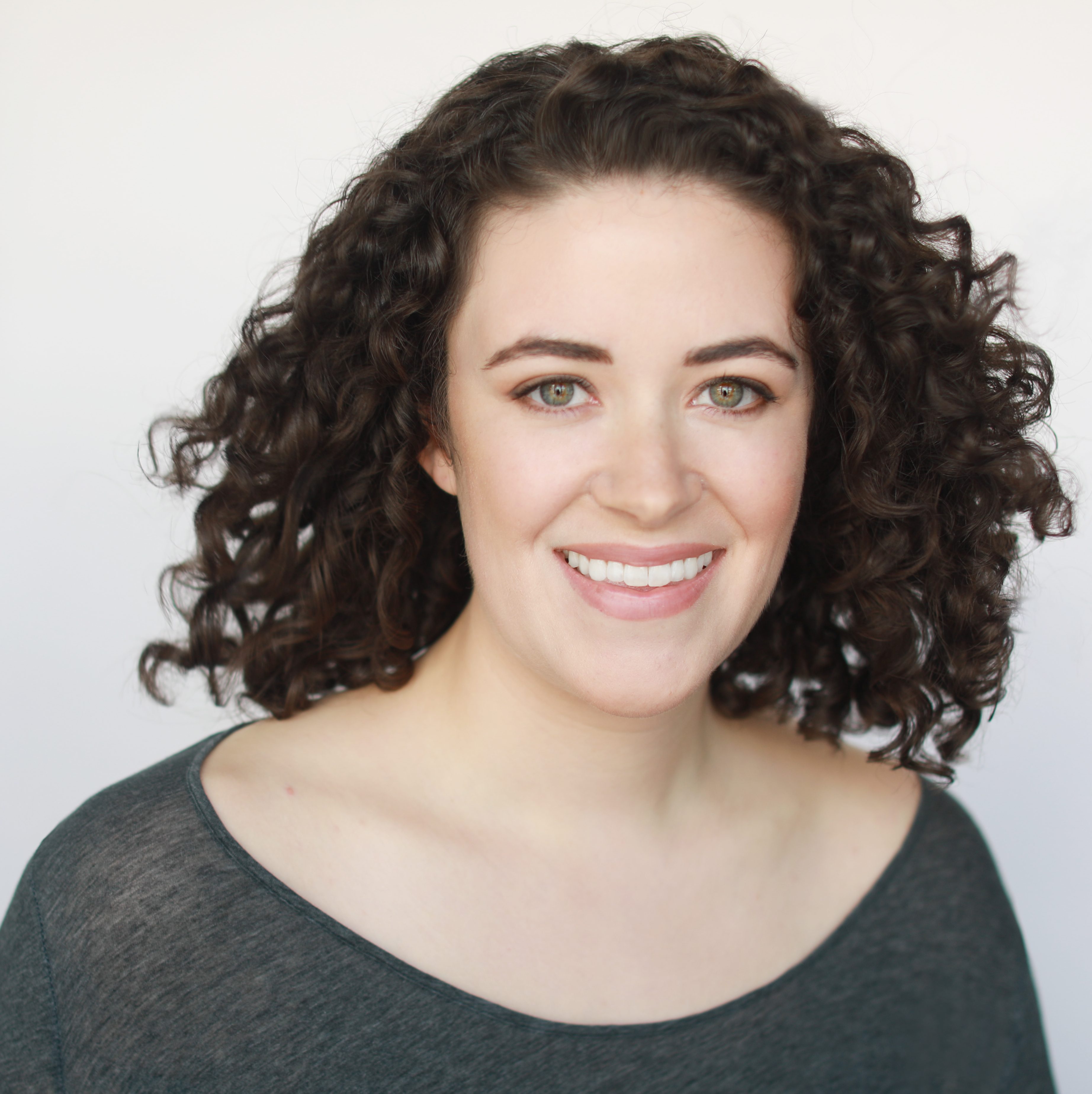 Carla is a white woman with shoulder-length dark brown curly hair and green eyes. She wears a dark gray scoop neck shirt and is smiling against a light gray background.