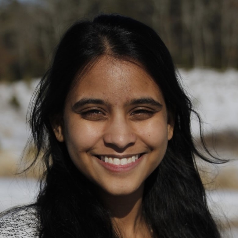 Leela, a South Asian femme, smiles widely with crescent eyes. Her long black hair wisps away from her face.