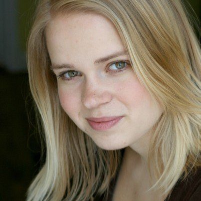 Maggie, ablonde, white woman with hazel eyes and a brown shirt is against a blurred background. She is in 3/4 profile to the camera with a slight smile on her face.